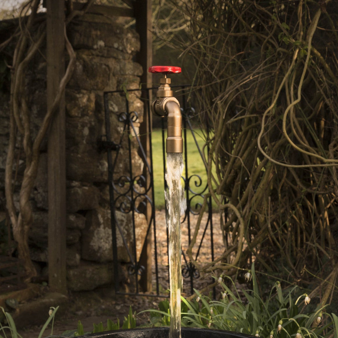 A floating tap water feature with a red gate valve tap attached to a black container, with water flowing into the container, set in a garden environment.