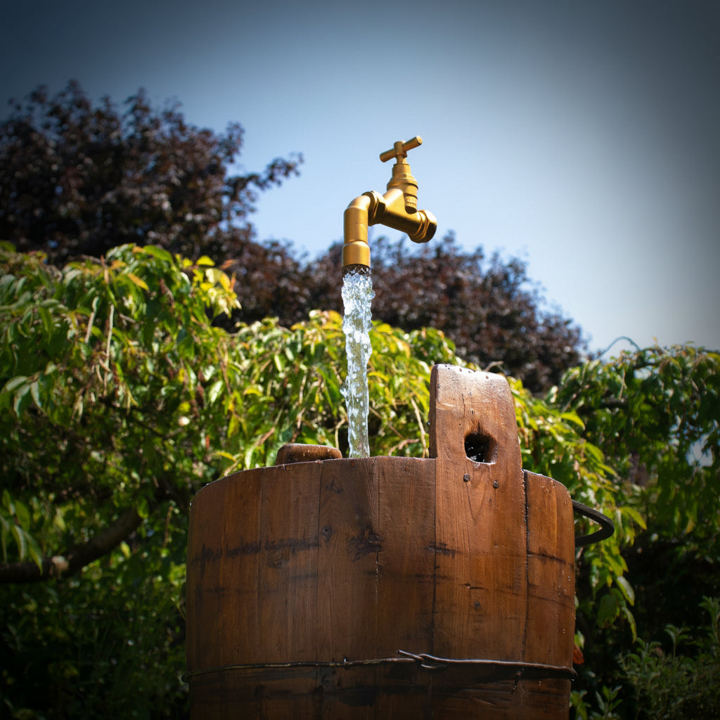 Gold faucet on a wooden barrel with water flowing, surrounded by greenery.