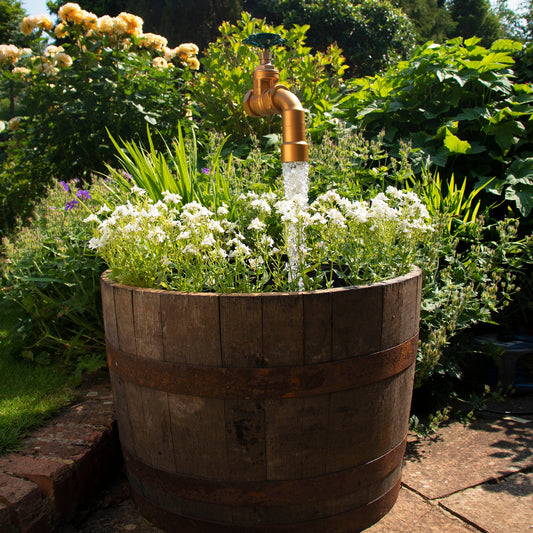 A half oak barrel water feature with a large brass tap installed in the center, surrounded by plants and set in an outdoor setting.