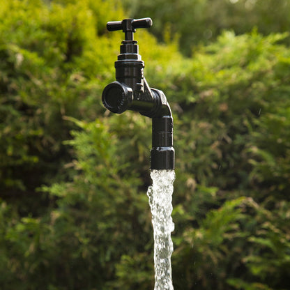 A floating tap water feature with a solid brass tap and soldered copper spout, appearing to float from the ground, with real running water, set in a garden environment with flowers in the background.
