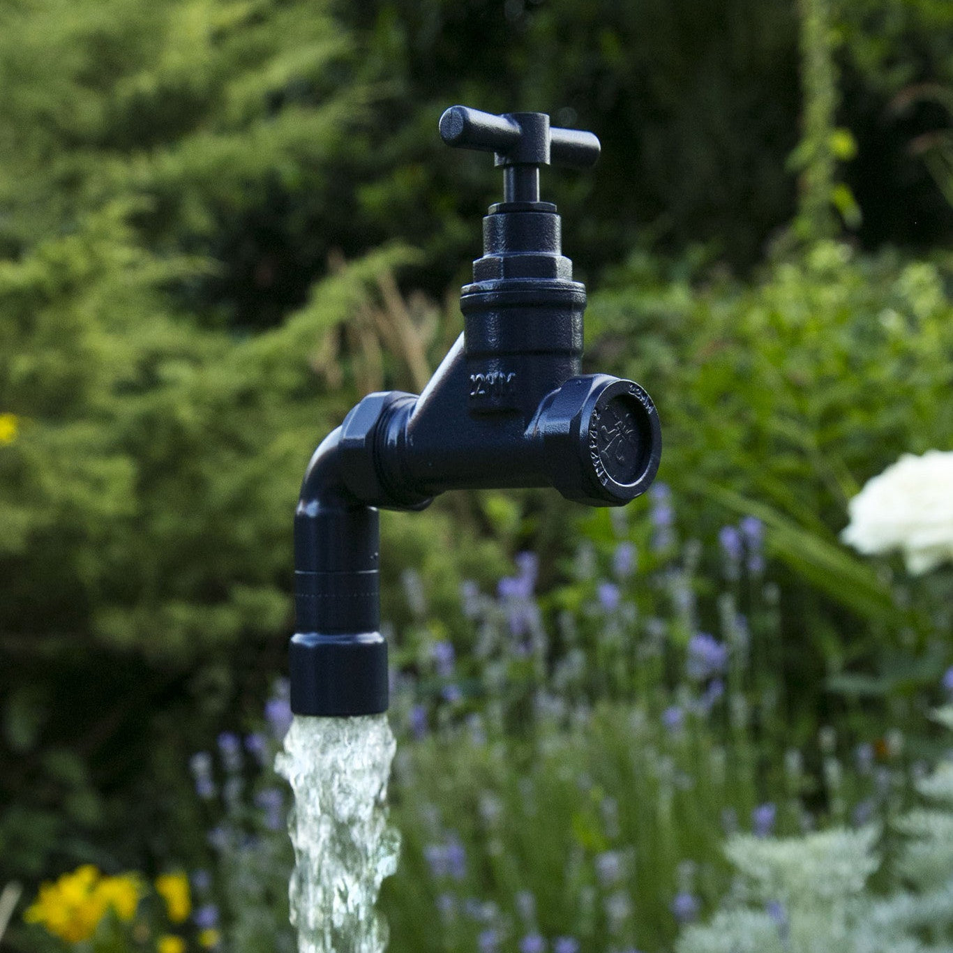 A floating tap water feature with a solid brass tap and soldered copper spout, appearing to float from the ground, with real running water, set in a garden environment with flowers in the background.