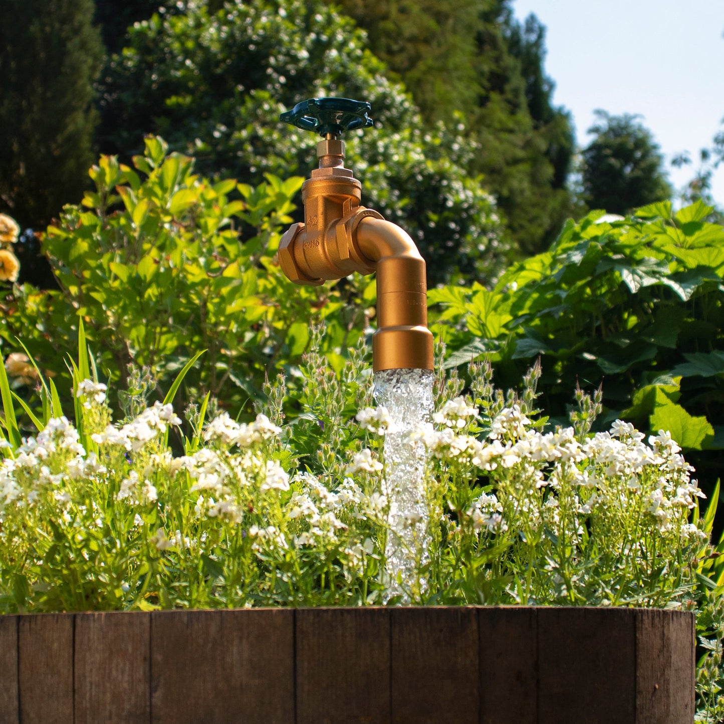 A half oak barrel water feature with a large brass tap installed in the center, surrounded by plants and set in an outdoor setting.