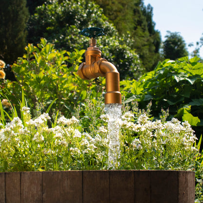 A half oak barrel water feature with a large brass tap installed in the center, surrounded by plants and set in an outdoor setting.