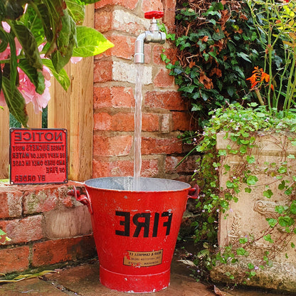 A floating tap water feature with a solid brass tap and soldered copper spout, running water, set in a garden environment with flowers in the background.