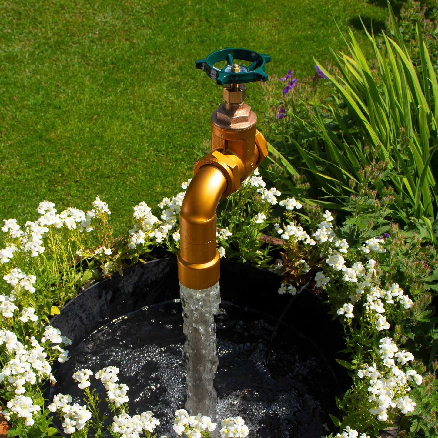 A half oak barrel water feature with a large brass tap installed in the center, surrounded by plants and set in an outdoor setting.