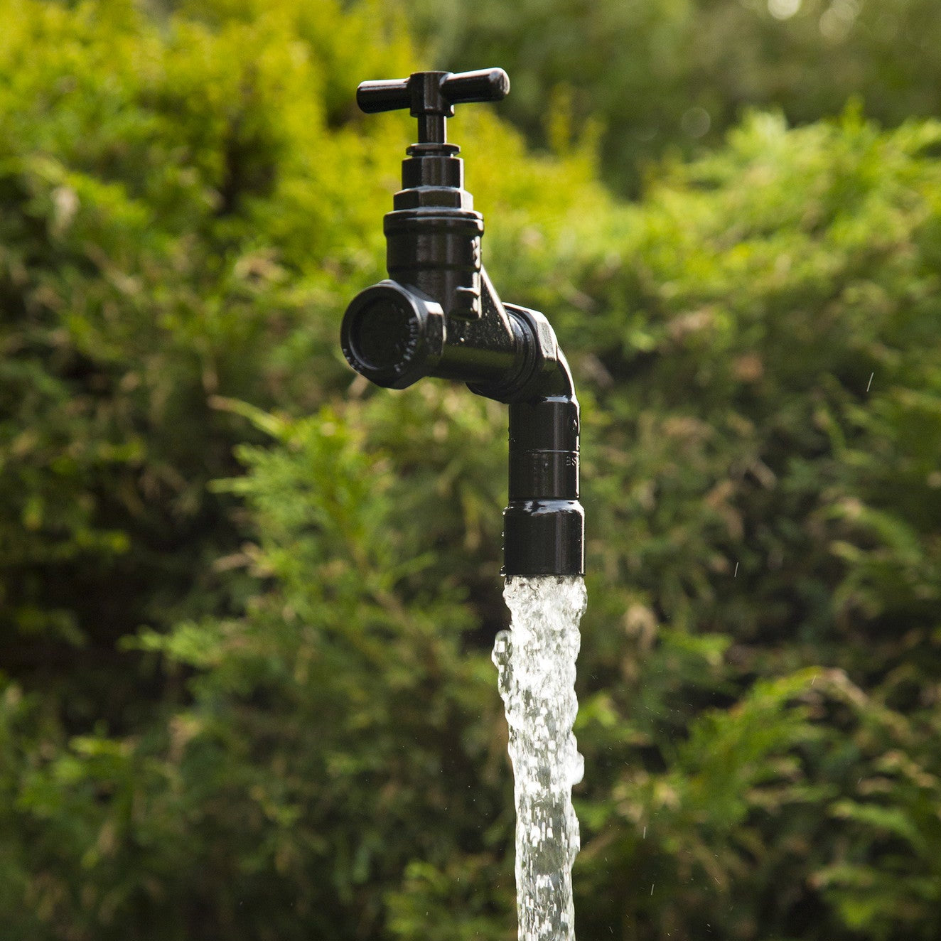 A floating tap water feature with a solid brass tap and soldered copper spout, running water, set in a garden environment with flowers in the background.