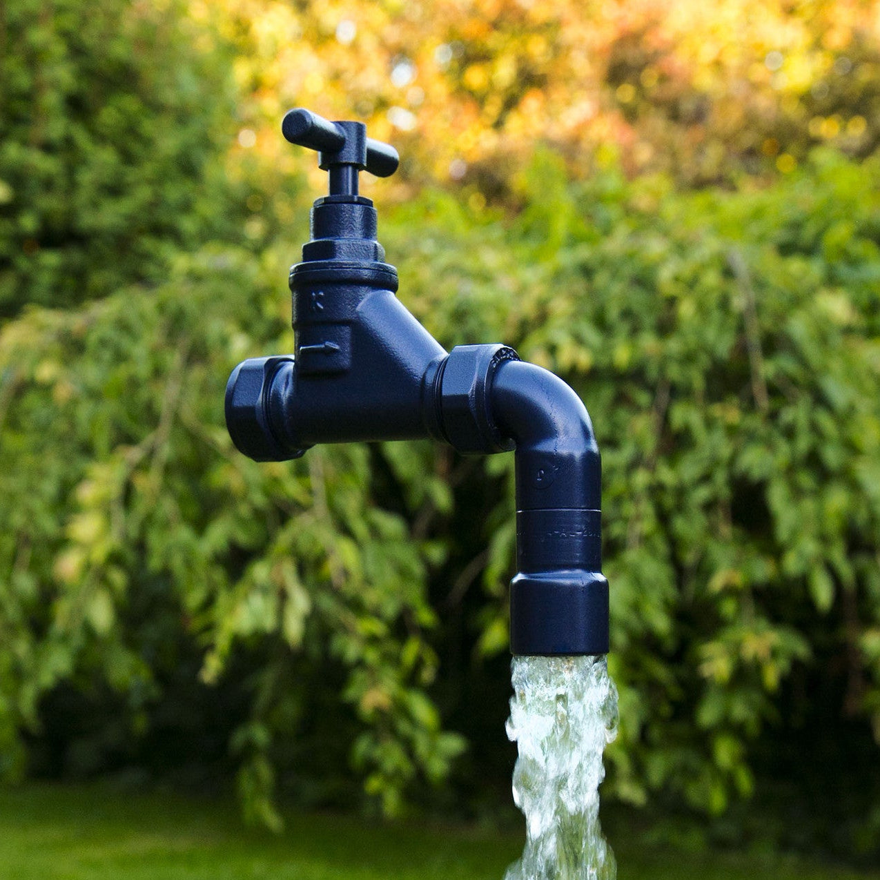 A floating tap water feature with a solid brass tap and soldered copper spout, running water, set in a garden environment with flowers in the background.