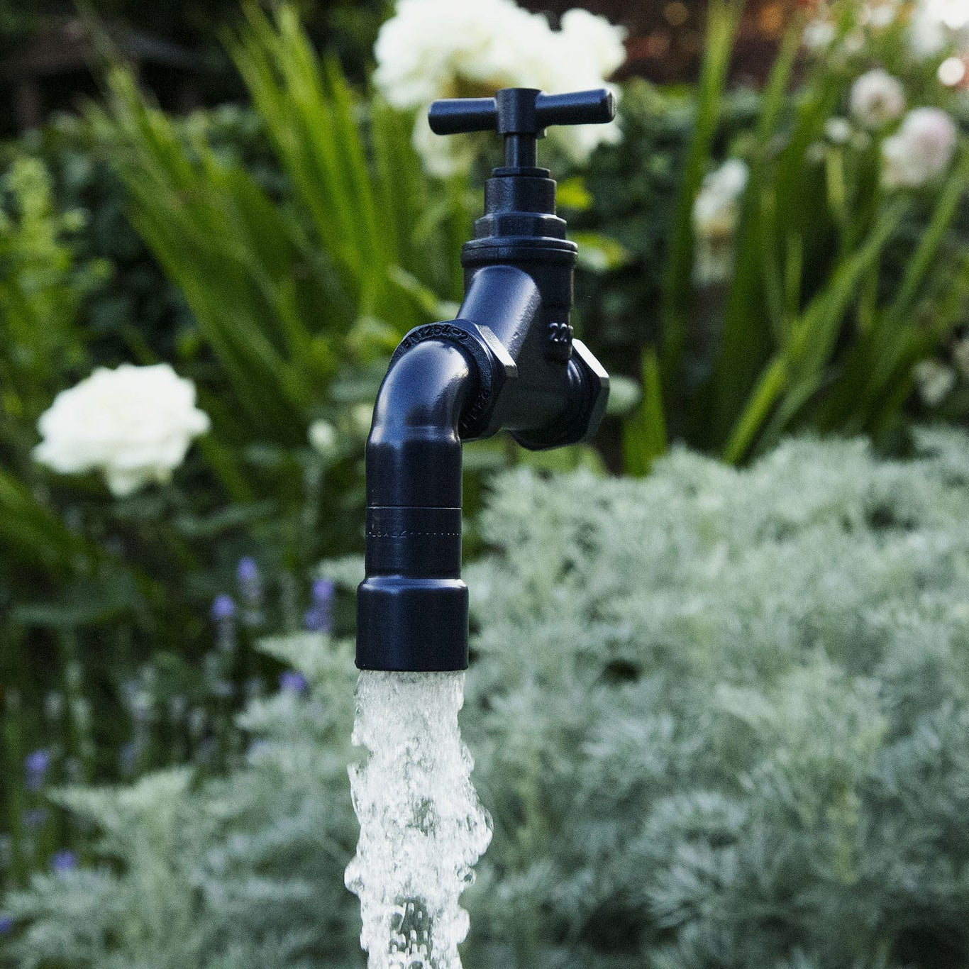 A floating tap water feature with a solid brass tap and soldered copper spout, running water, set in a garden environment with flowers in the background.