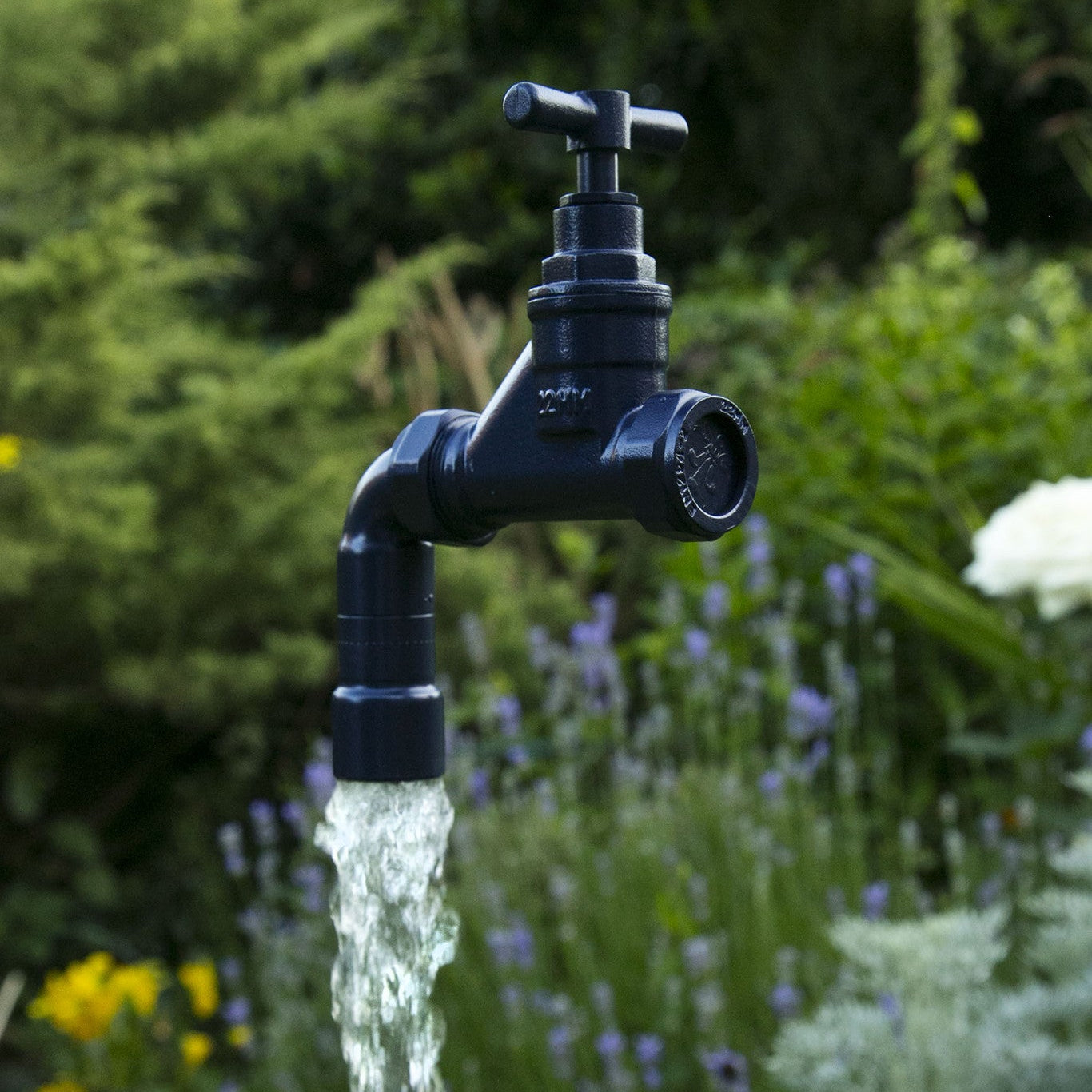 A floating tap water feature with a solid brass tap and soldered copper spout, running water, set in a garden environment with flowers in the background.