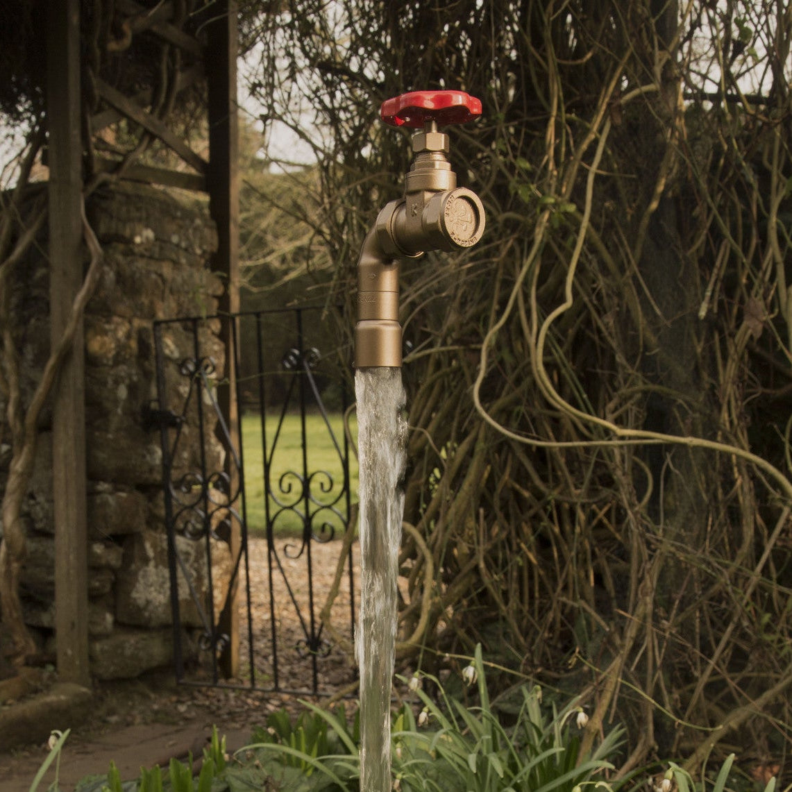 Bronze faucet with red handle dripping water outdoors near a stone wall and metal gate.