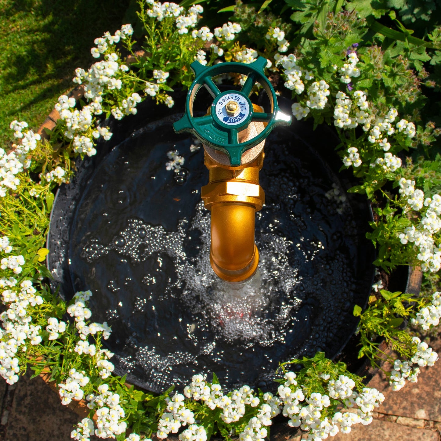 A half oak barrel water feature with a large brass tap installed in the center, surrounded by plants and set in an outdoor setting.