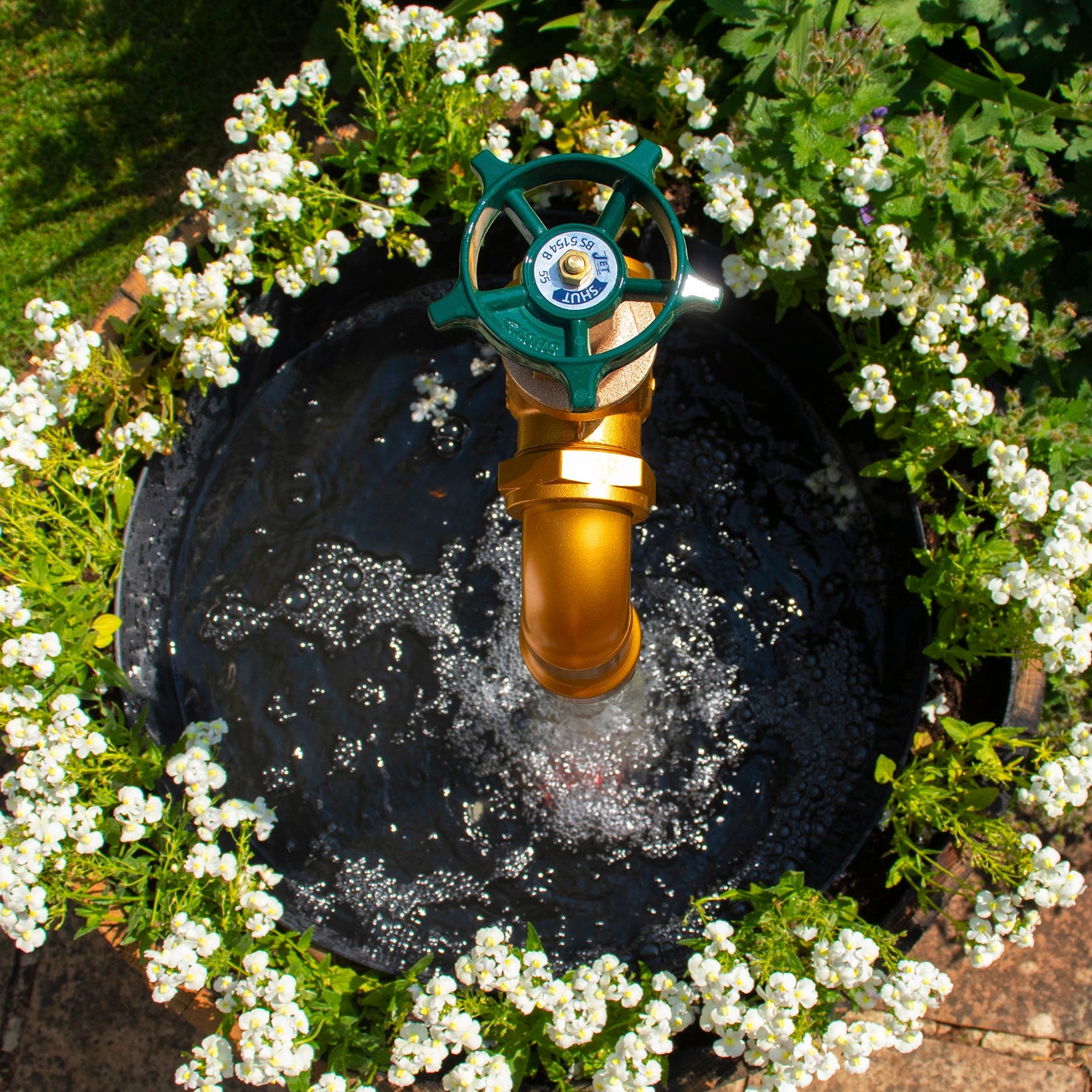 A half oak barrel water feature with a large brass tap installed in the center, surrounded by plants and set in an outdoor setting.