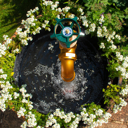 A half oak barrel water feature with a large brass tap installed in the center, surrounded by plants and set in an outdoor setting.