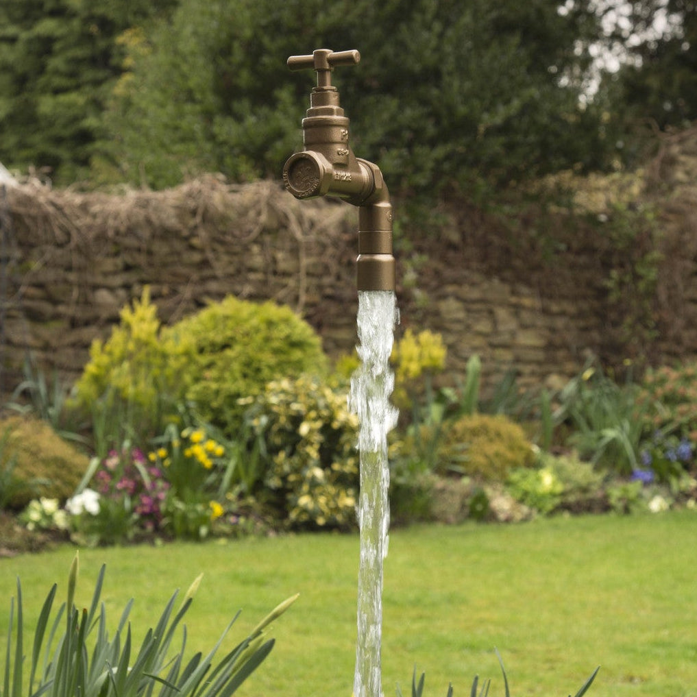 A Golden floating tap water feature with a solid brass tap and soldered copper spout, running water, set in a garden environment with flowers and shrubs in the background.