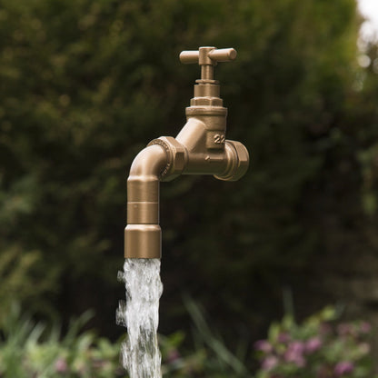 A floating tap water feature with a solid brass tap and soldered copper spout, running water, set in a garden environment with flowers in the background.