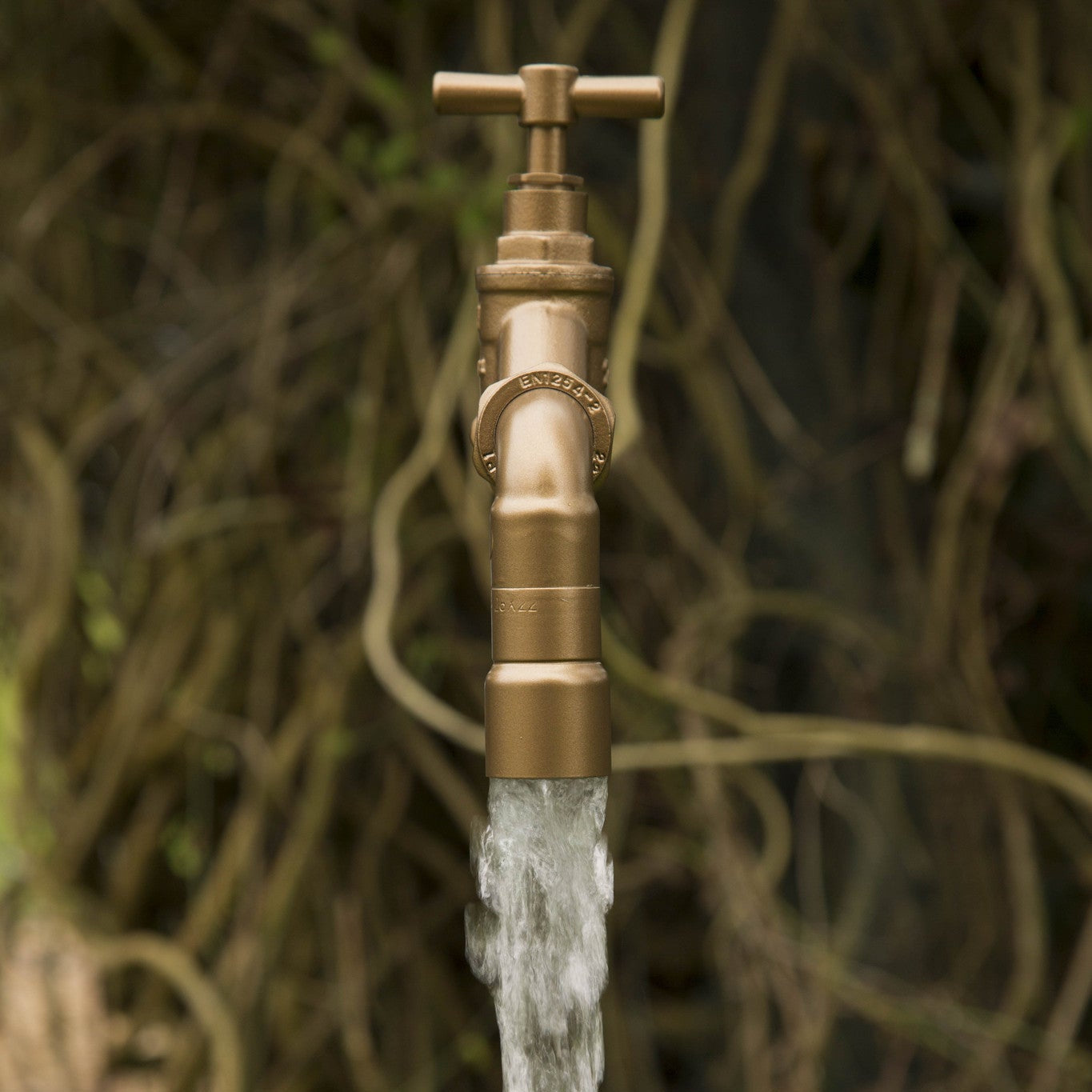 A floating tap water feature with a solid brass tap and soldered copper spout, running water, set in a garden environment with flowers in the background.