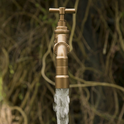 A floating tap water feature with a solid brass tap and soldered copper spout, running water, set in a garden environment with flowers in the background.