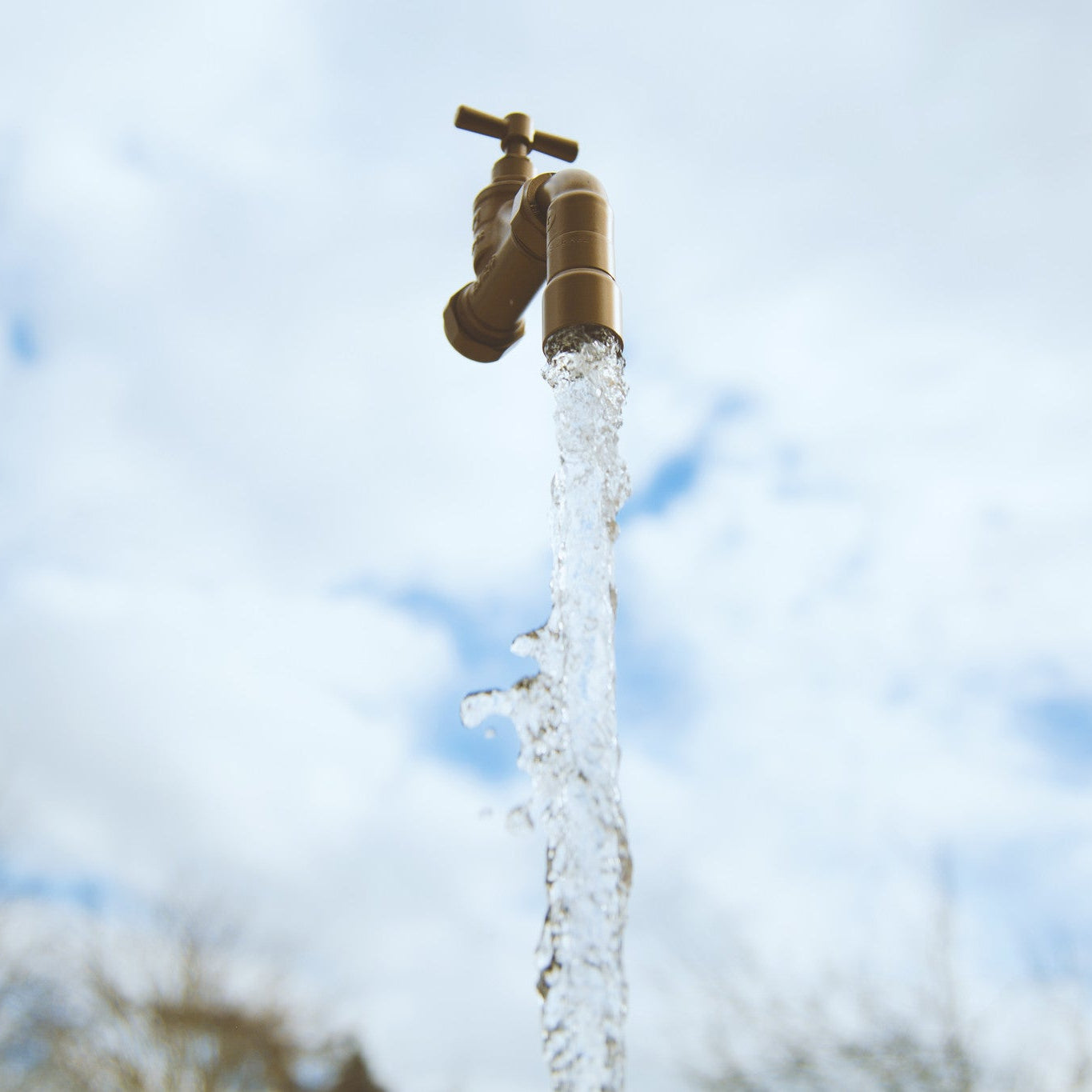 A floating tap water feature with a solid brass tap and soldered copper spout, running water, set in a garden environment with flowers in the background.