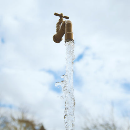 A floating tap water feature with a solid brass tap and soldered copper spout, running water, set in a garden environment with flowers in the background.
