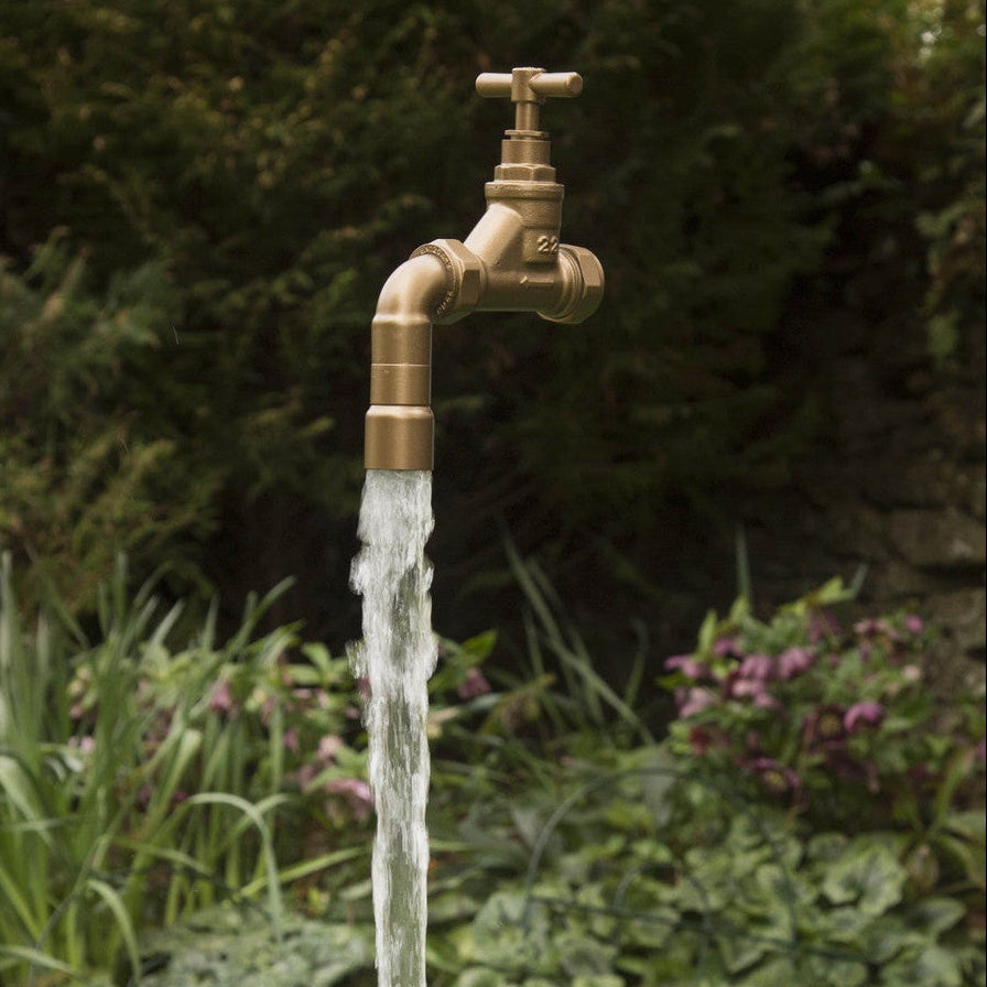 A floating tap water feature with a solid brass tap and soldered copper spout, running water, set in a garden environment with flowers in the background.