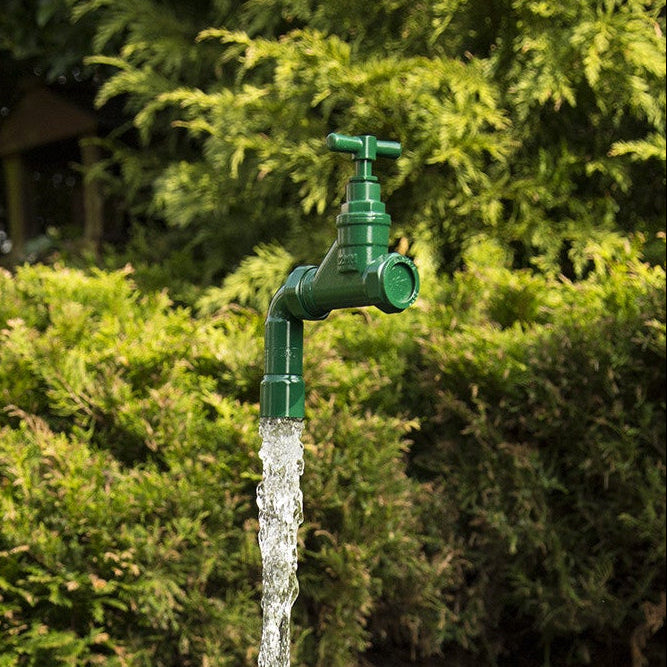 A floating tap water feature with a solid brass tap and soldered copper spout, running water, set in a garden environment with flowers in the background.