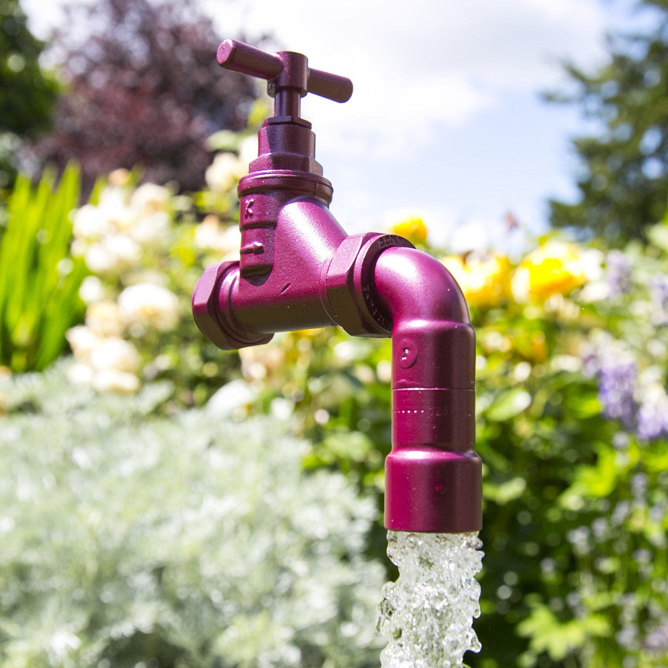 A floating tap water feature with a solid brass tap and soldered copper spout, running water, set in a garden environment with flowers in the background.