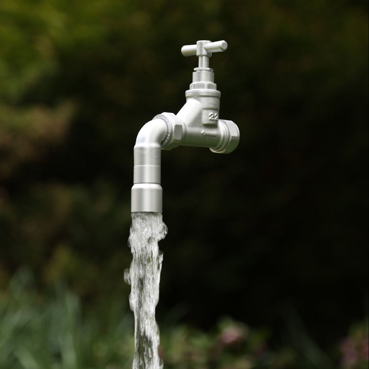 A Silver floating tap water feature with a solid brass tap and soldered copper spout, running water, set in a garden environment with plants in the background.