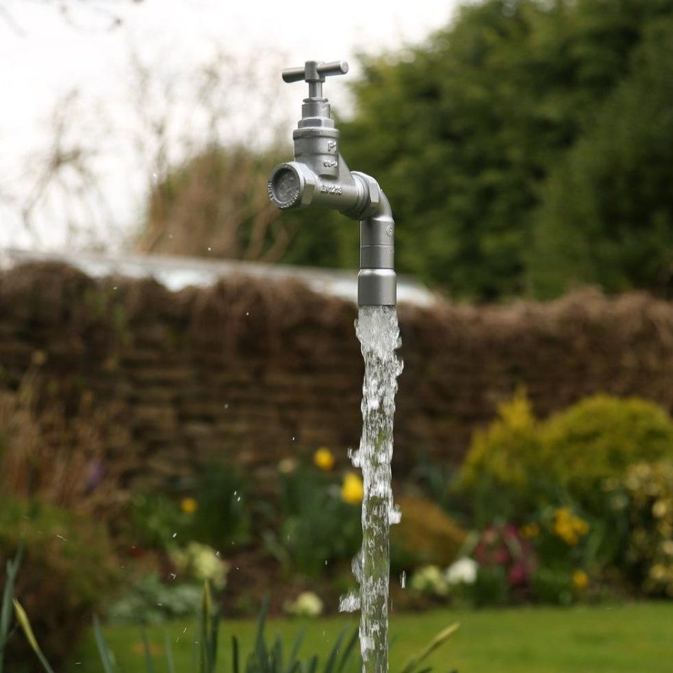 A floating tap water feature with a solid brass tap and soldered copper spout, running water, set in a garden environment with flowers in the background.