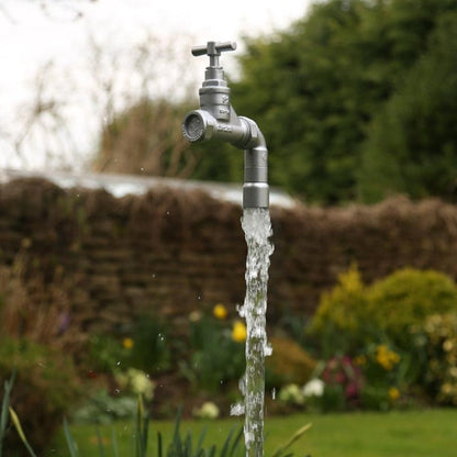 A floating tap water feature with a solid brass tap and soldered copper spout, running water, set in a garden environment with flowers in the background.