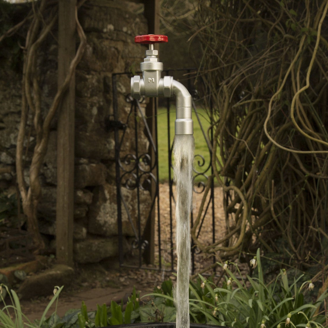 A floating tap water feature with a red gate valve tap attached to a black container, with water flowing into the container, set in a garden environment.