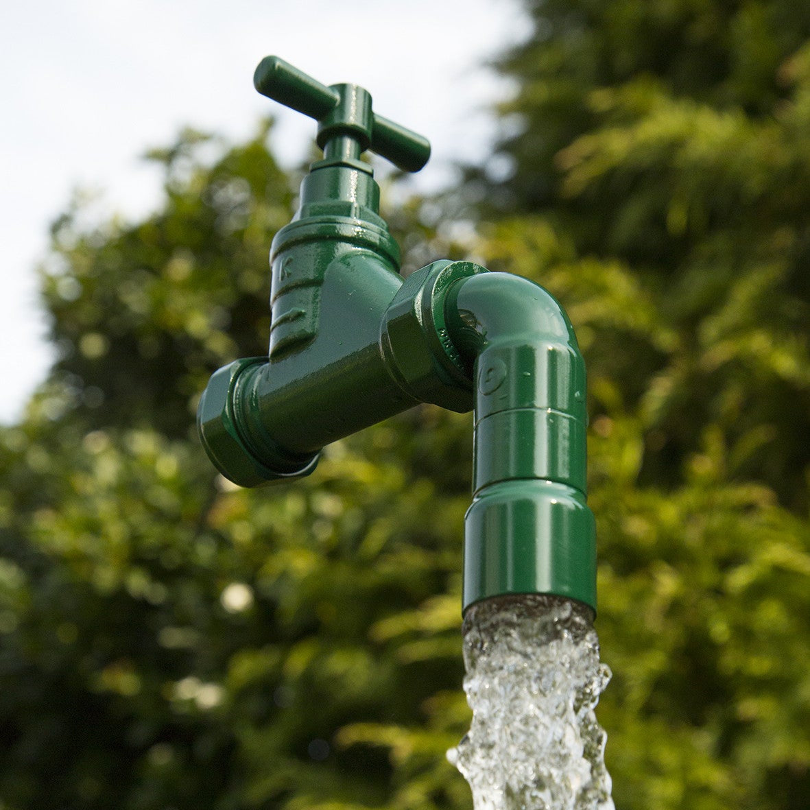 A floating tap water feature with a solid brass tap and soldered copper spout, appearing to float from the ground, with real running water, set in a garden environment with flowers in the background.
