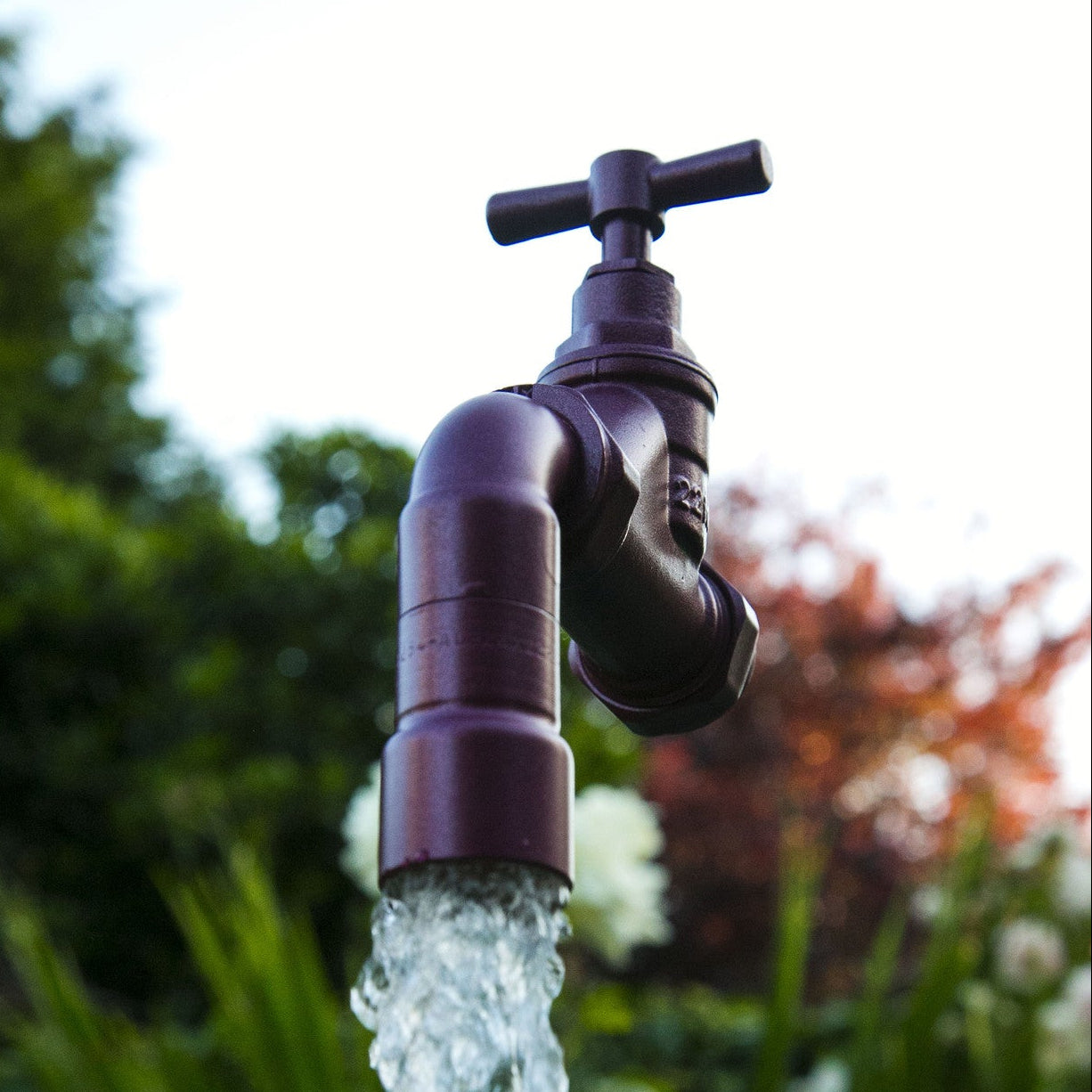 A floating tap water feature with a solid brass tap and soldered copper spout, appearing to float from the ground, with real running water, set in a garden environment with flowers in the background.