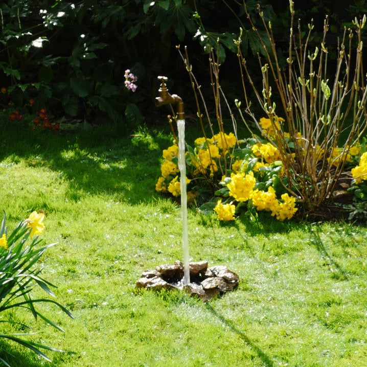 A floating tap water feature with a solid brass tap and soldered copper spout, appearing to float from the ground, with real running water, set in a garden environment with flowers in the background.
