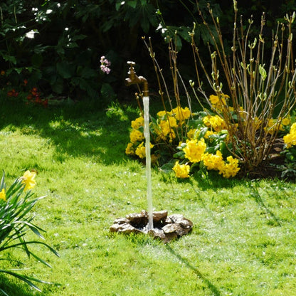 A floating tap water feature with a solid brass tap and soldered copper spout, appearing to float from the ground, with real running water, set in a garden environment with flowers in the background.