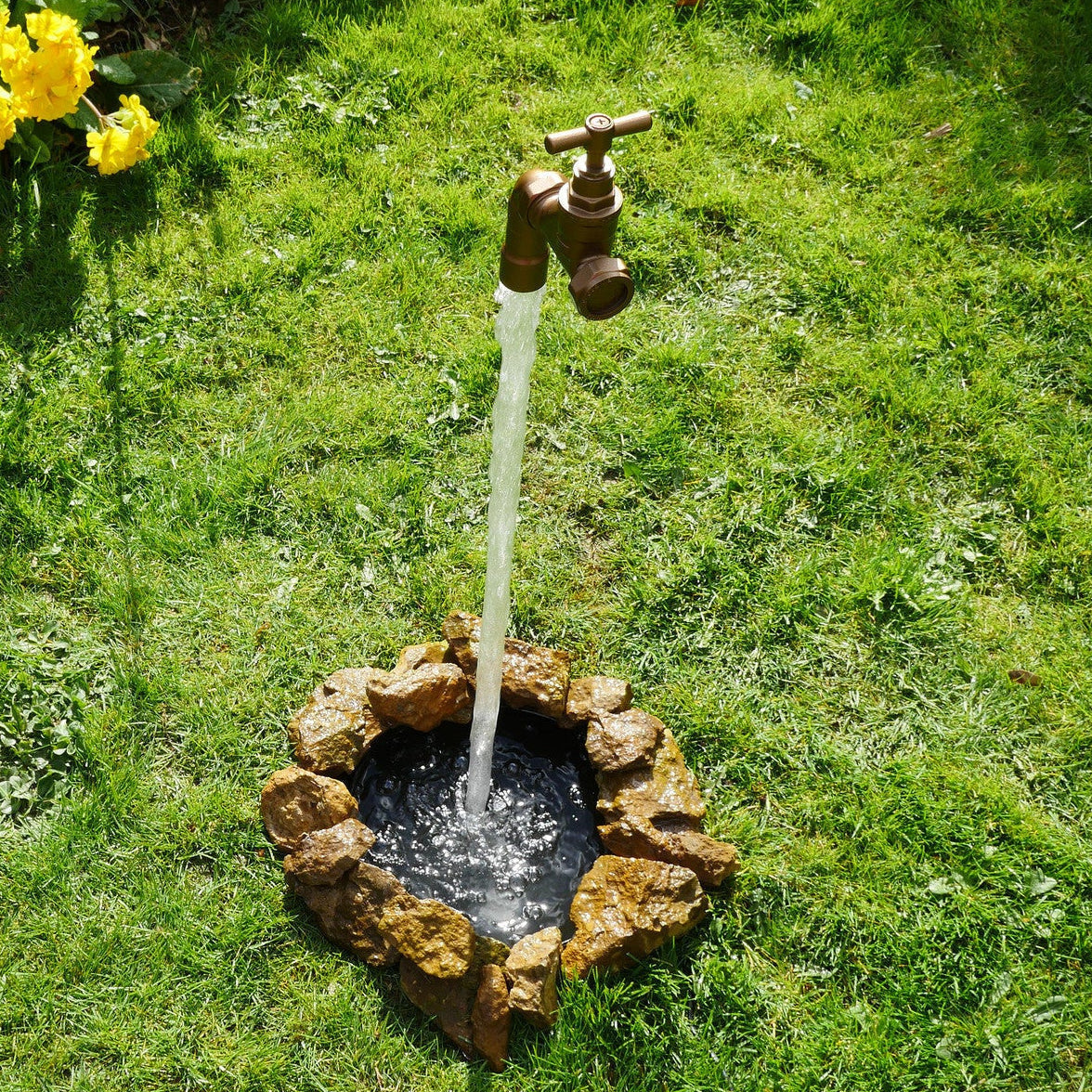 A floating tap water feature with a solid brass tap and soldered copper spout, appearing to float from the ground, with real running water, set in a garden environment with flowers in the background.