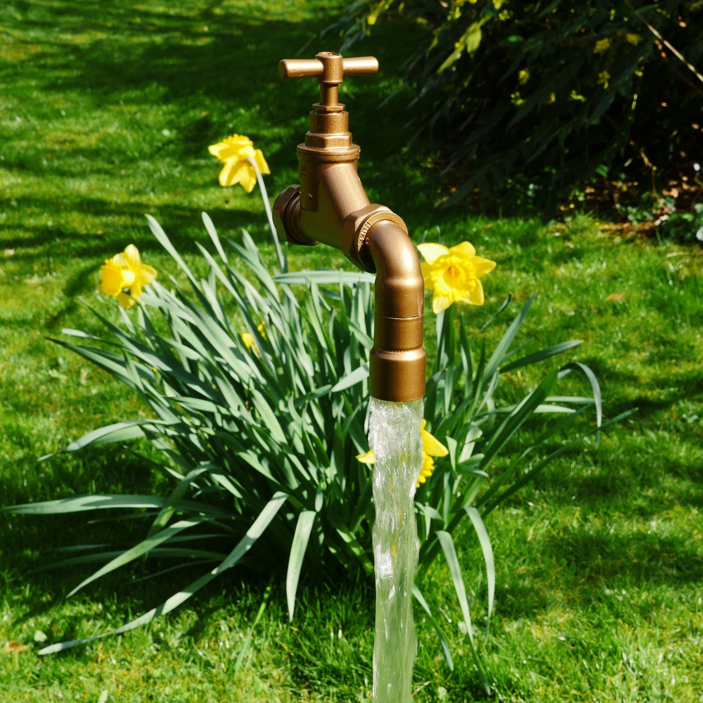 A floating tap water feature with a solid brass tap and soldered copper spout, appearing to float from the ground, with real running water, set in a garden environment with flowers in the background.