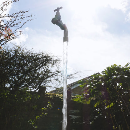 A floating tap water feature with a solid brass tap and soldered copper spout, appearing to float from the ground, with real running water, set in a garden environment with flowers in the background.