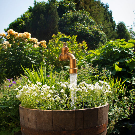 A large floating tap installed on top of a barrel surrounded by plants, with a clear sky and trees in the background.